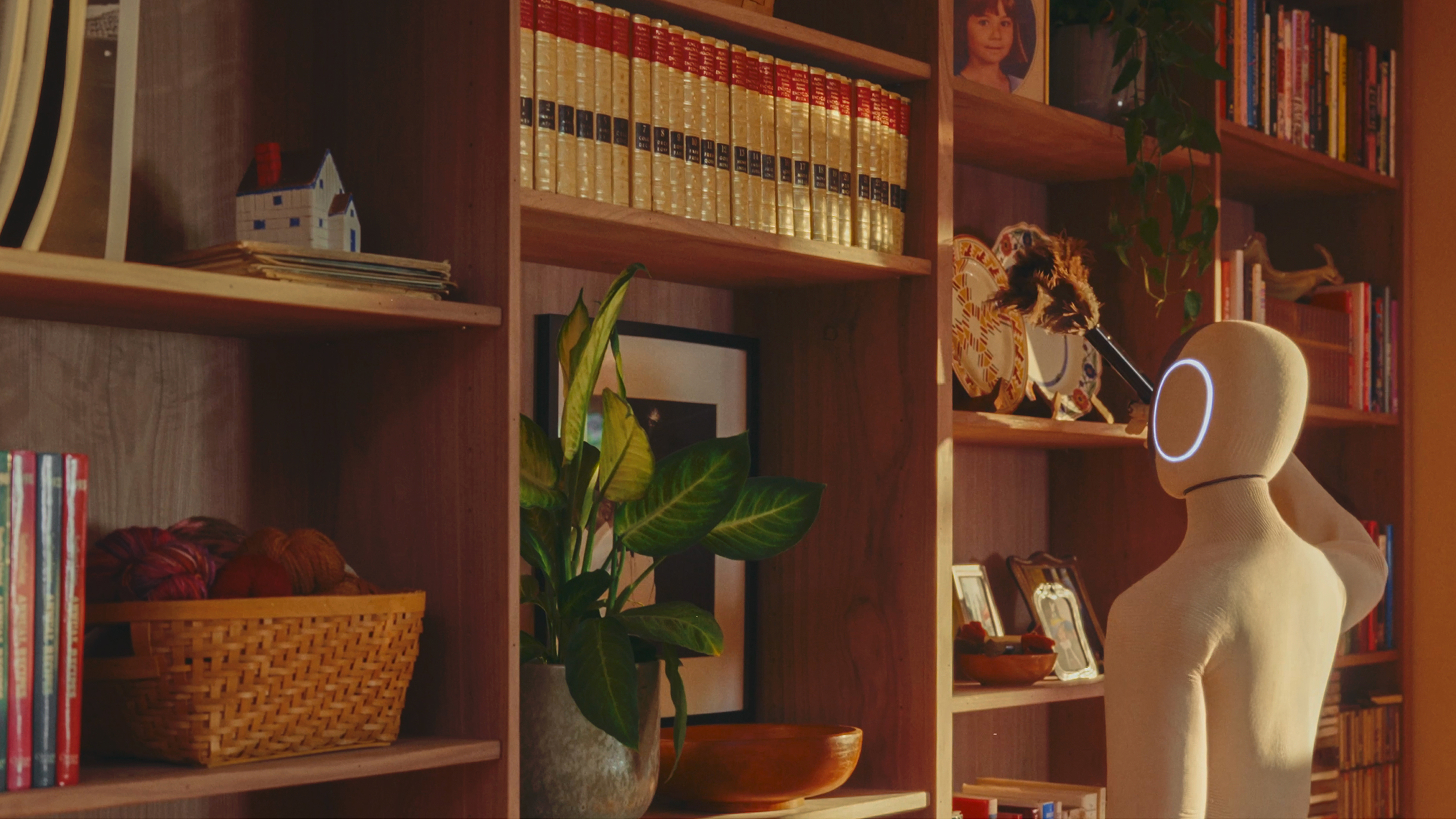 A knit-covered humanoid robot holds a feather duster up to a wooden bookshelf in a home library.
