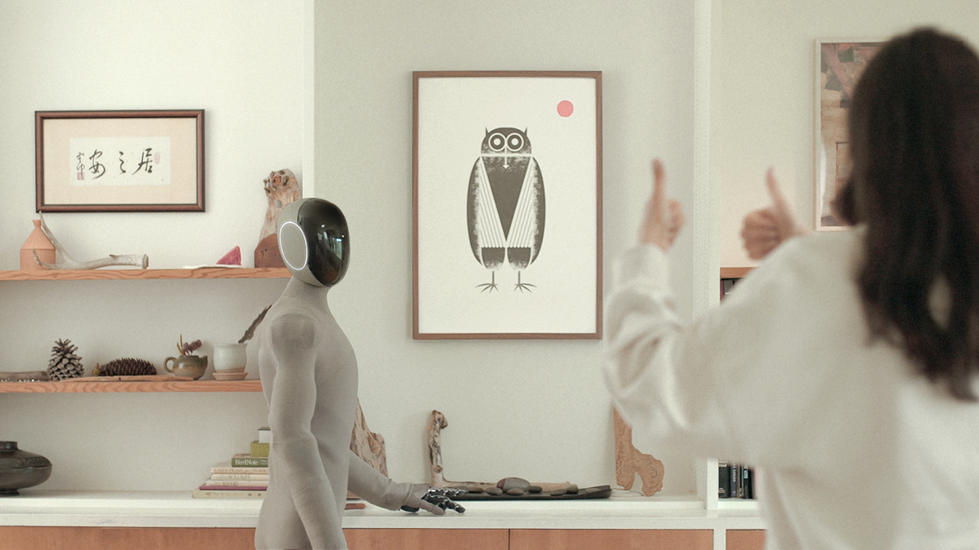 A knit-covered humanoid robot with a dark ring-faced head stands in a room as a person raises two thumbs-up nearby.