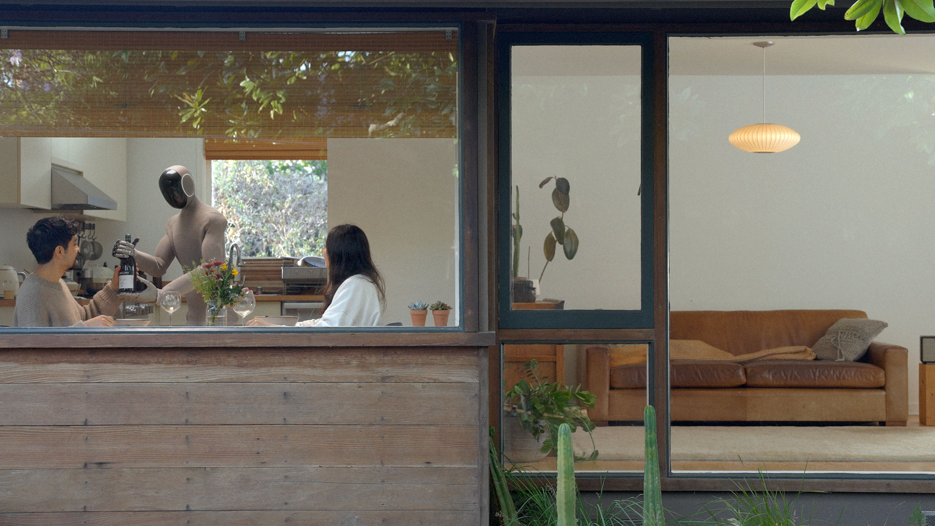 Seen through a window, a knit-covered humanoid robot stands at a kitchen counter near two seated people in a home.