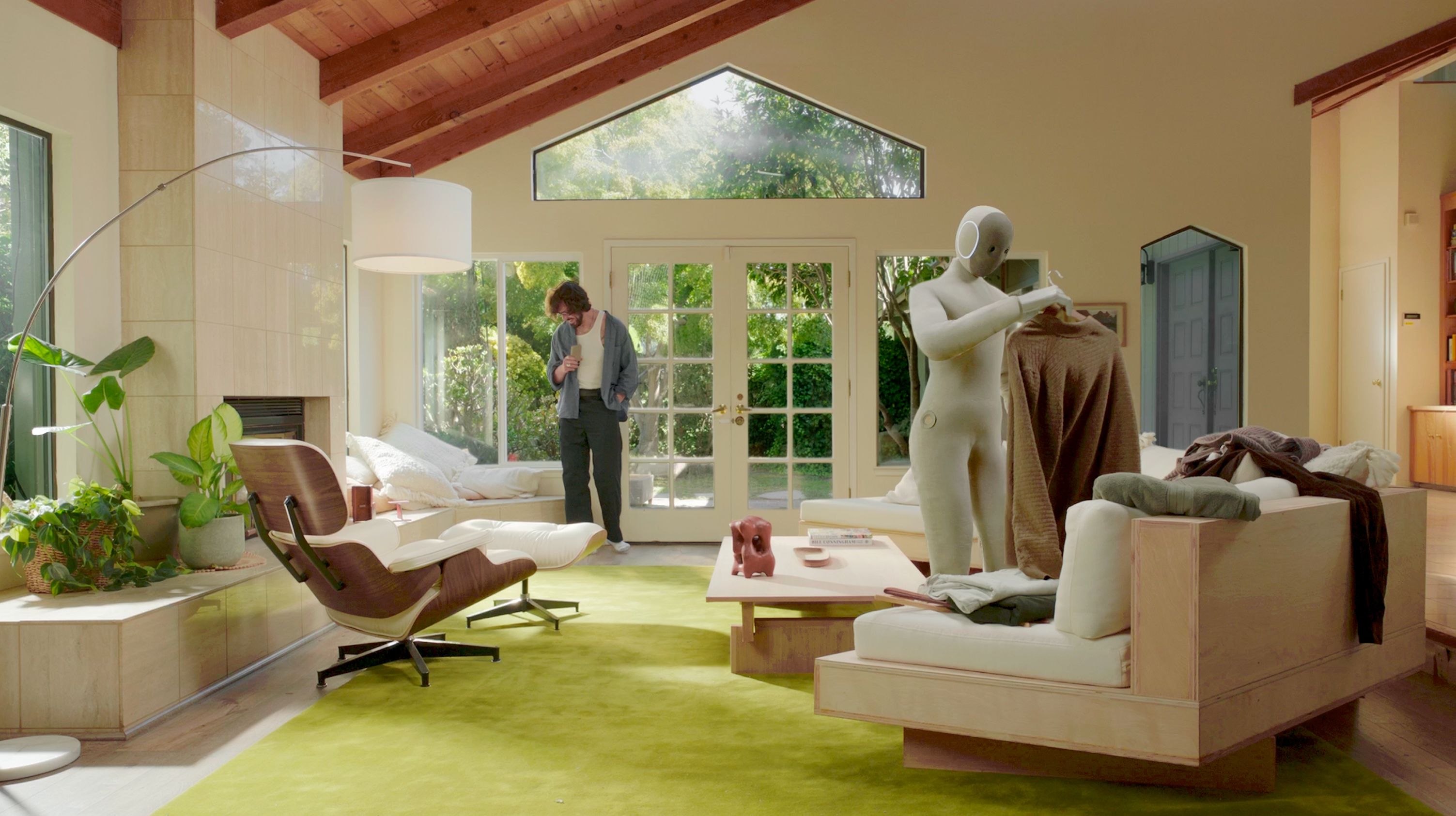 A knit-covered humanoid robot holds a draped garment over one arm in a sunlit living room while a person stands by glass doors.