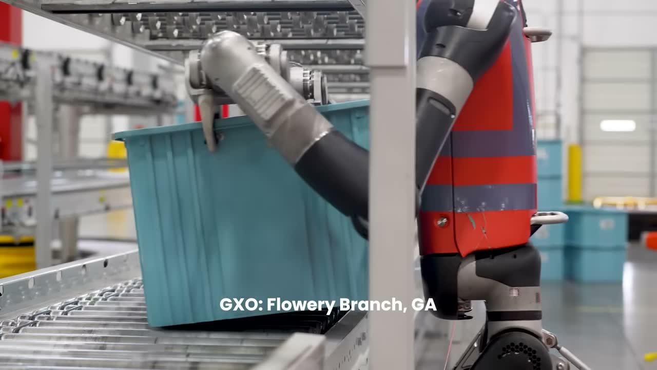 Agility Robotics Digit humanoid robot moving a tote onto a conveyor in a warehouse. Still from the official Agility Robotics video "Digit's First Day of Work at GXO" (2024); the frame carries the video's own on-screen caption.