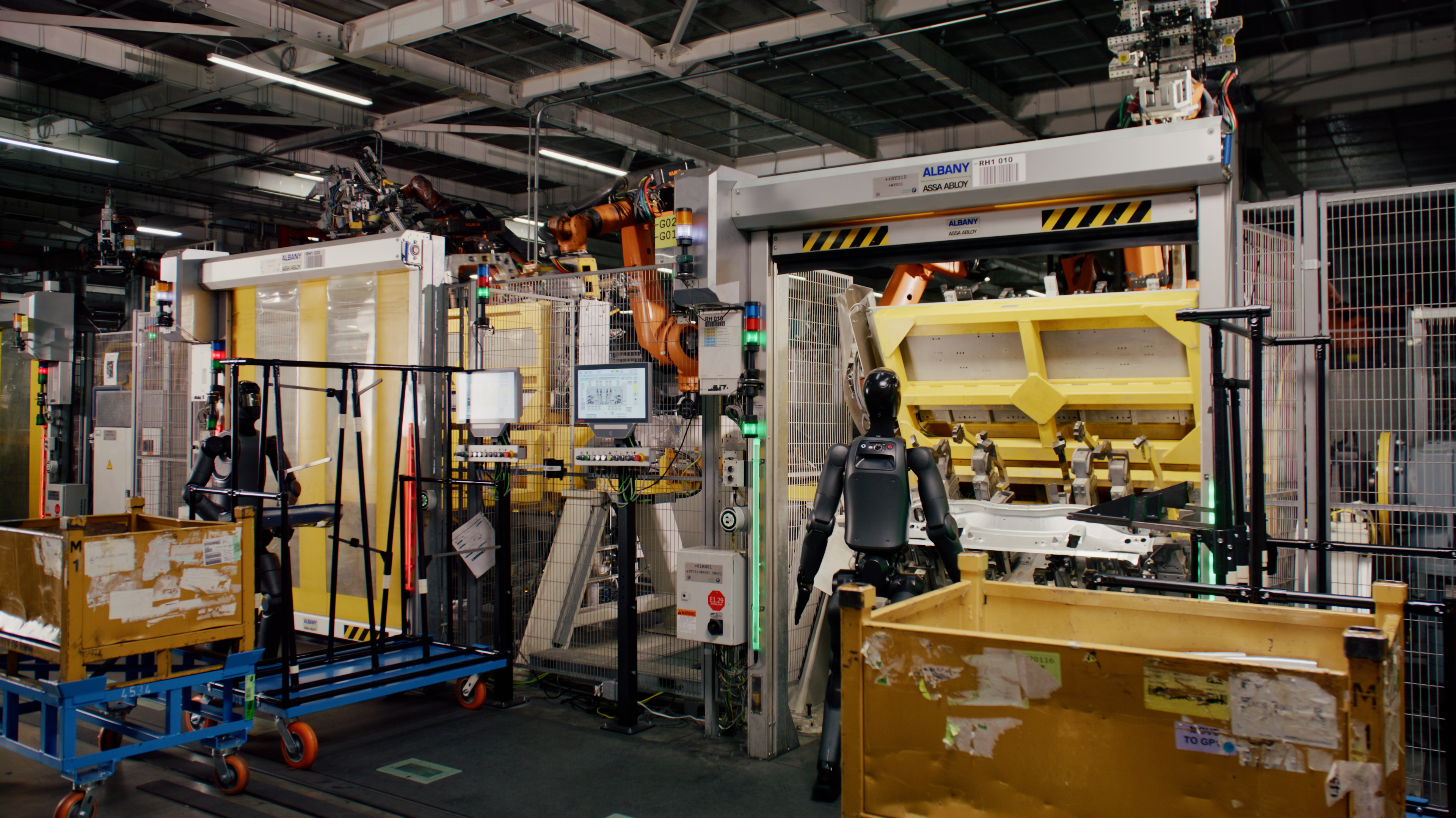 Two black Figure 02 humanoid robots at workstations on an automotive body-shop line with orange industrial robots and safety gates.