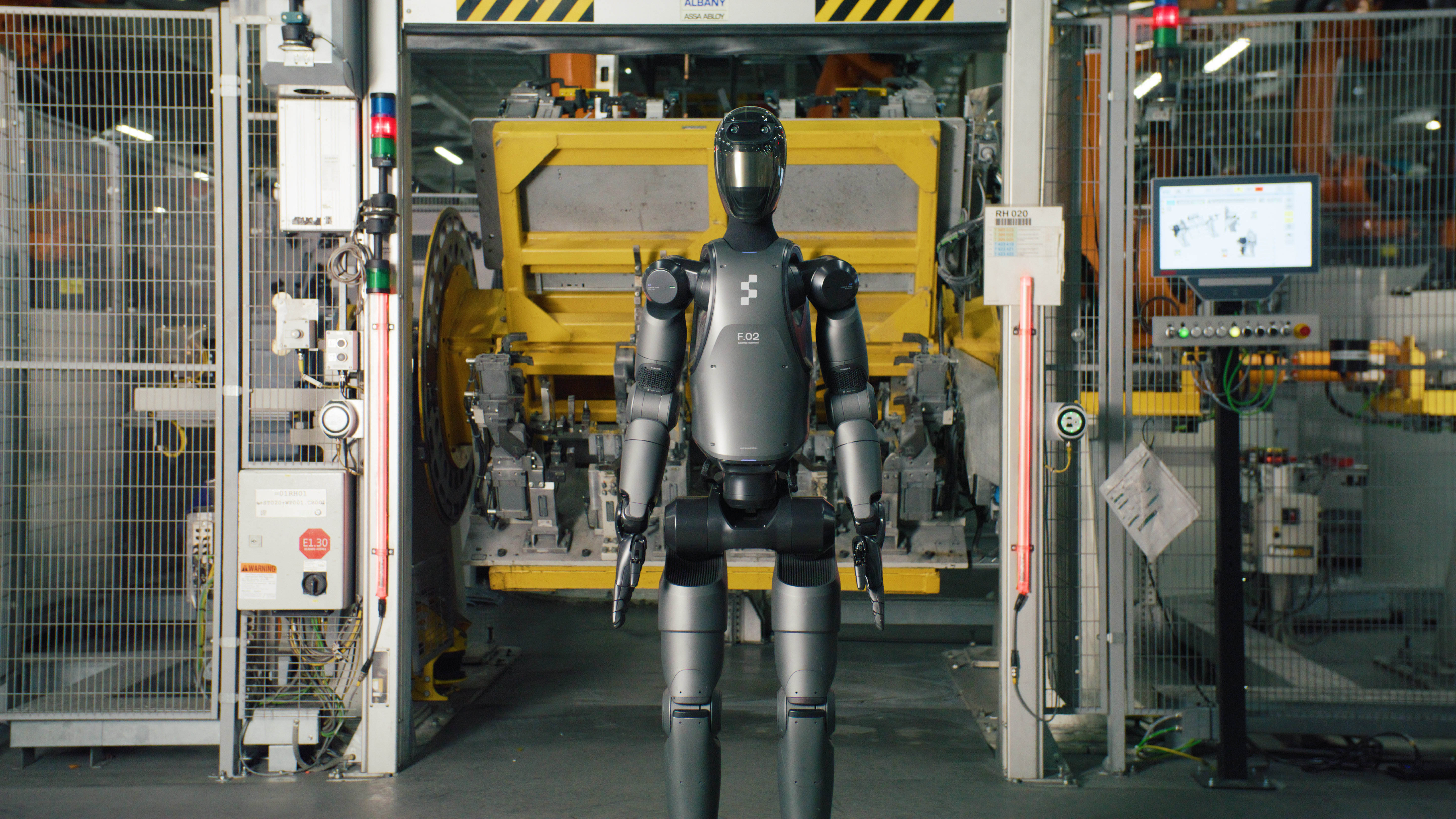 A black Figure 02 humanoid robot standing front-facing at an automotive body-shop station, with 'F.02' and the Figure logo on its chest.