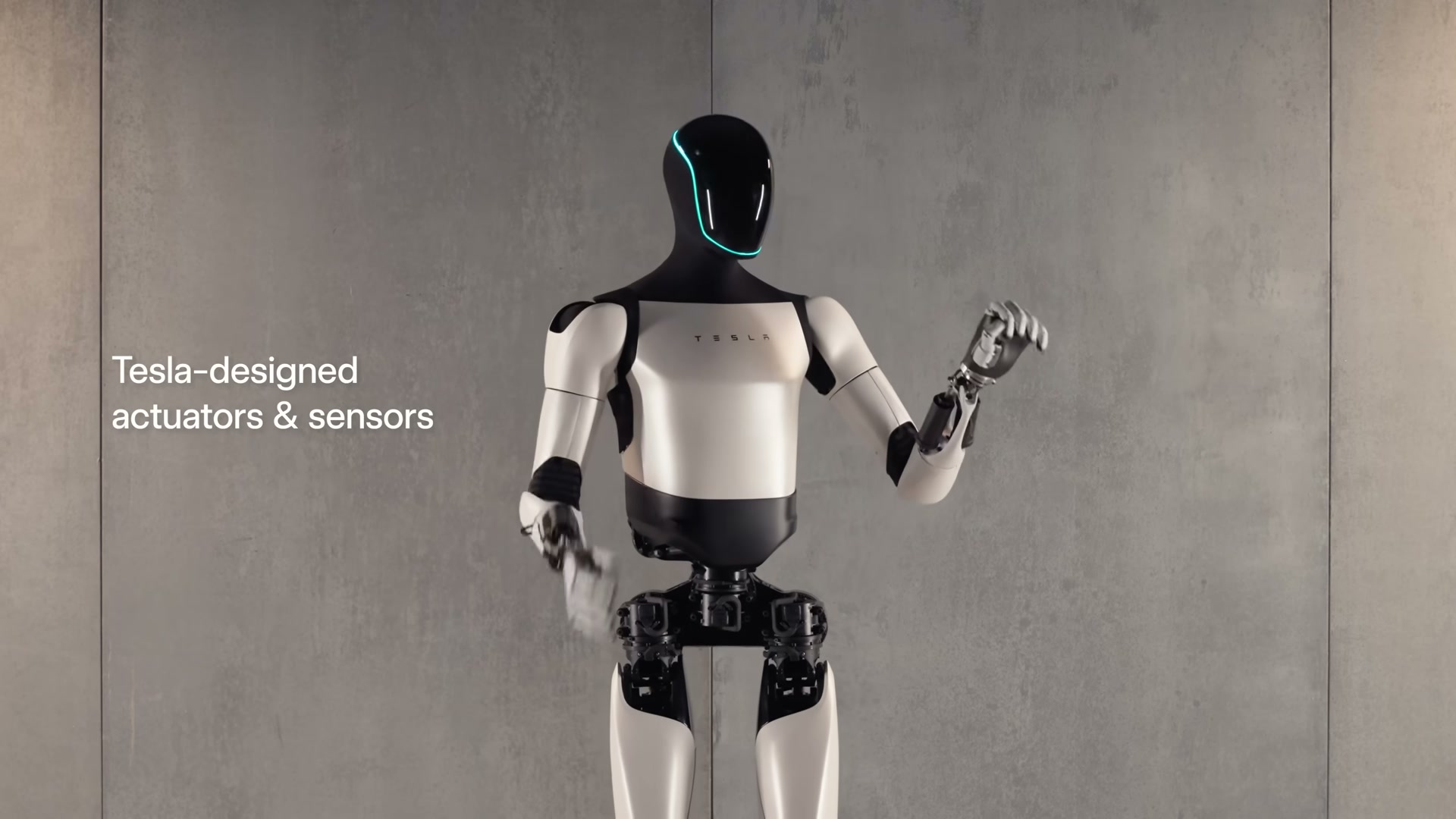 A Tesla Optimus (Gen 2) robot standing in a three-quarter pose against a plain concrete studio wall, one hand raised, with the on-screen caption 'Tesla-designed actuators & sensors'.