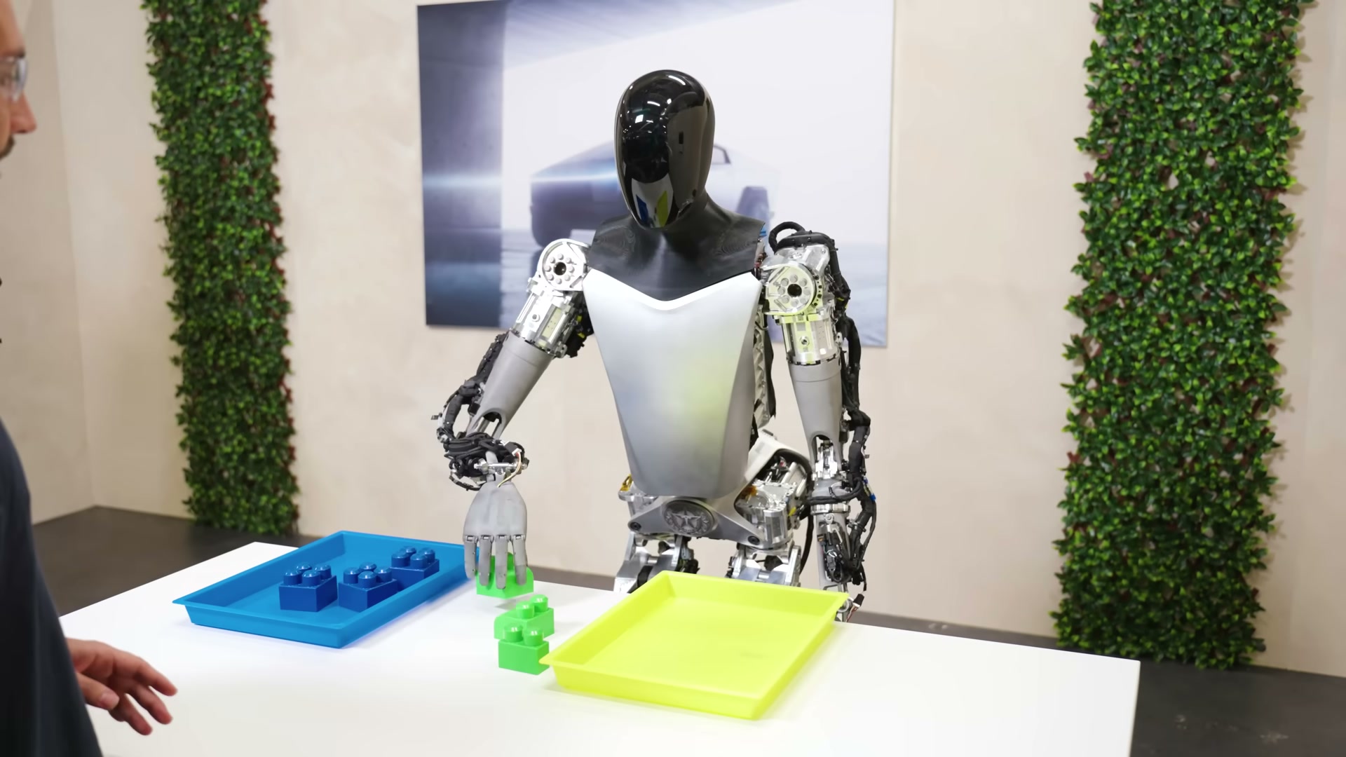 A Tesla Optimus robot with exposed silver internals standing behind a white studio table, its open right hand hovering over a cluster of green toy blocks between a blue tray of blue blocks and an empty yellow tray, with a person's hand resting on the near edge of the table and a Cybertruck poster behind.