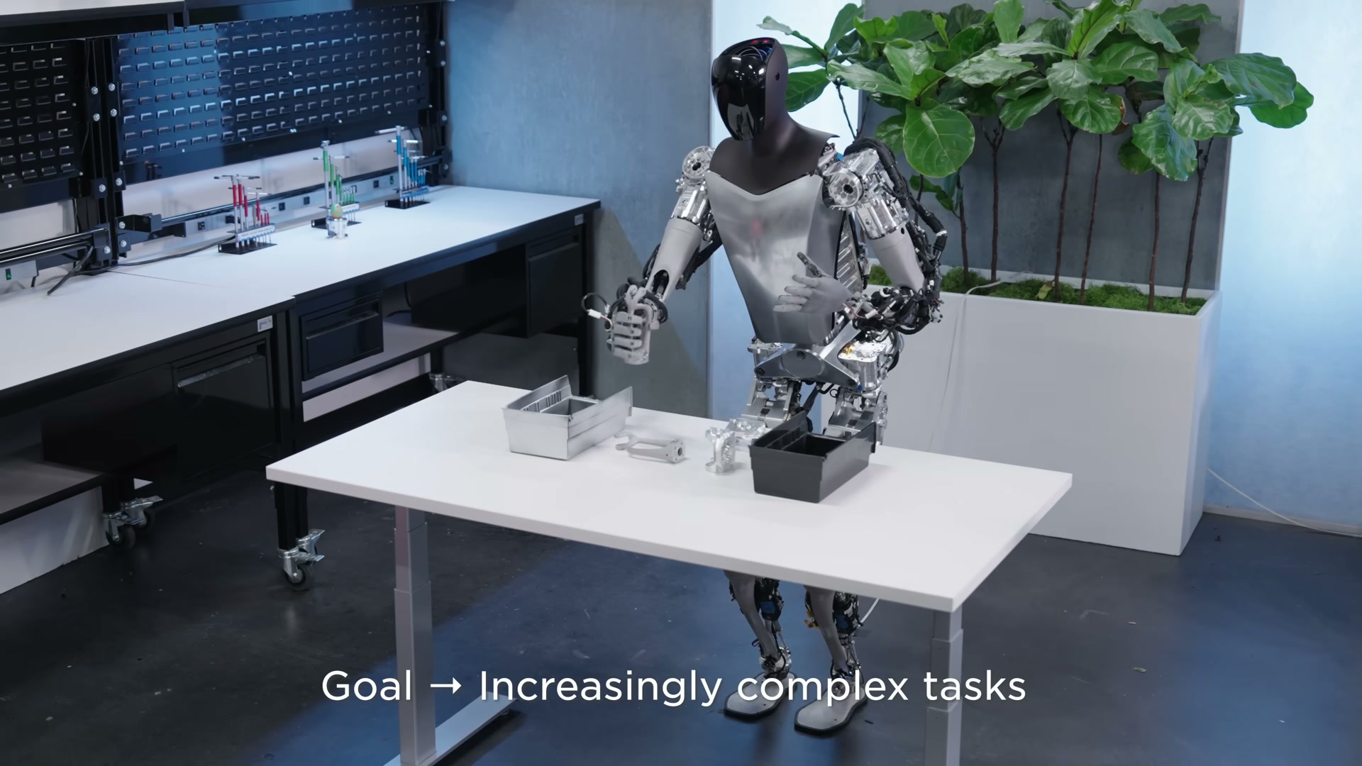 A development Tesla Optimus robot with exposed silver actuators standing at a white workbench that holds two metal bins and several machined metal parts, one hand extended toward the parts, with a pegboard tool wall and a fiddle-leaf plant behind and an on-screen caption about performing increasingly complex tasks.