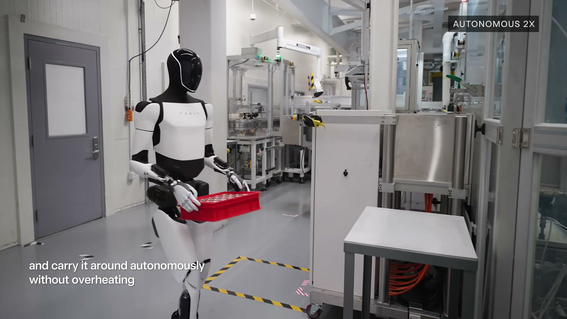 A production-design Tesla Optimus robot carrying a red plastic bin of small parts in both hands as it walks through a factory corridor lined with manufacturing equipment, with an 'AUTONOMOUS 2X' label in the corner.