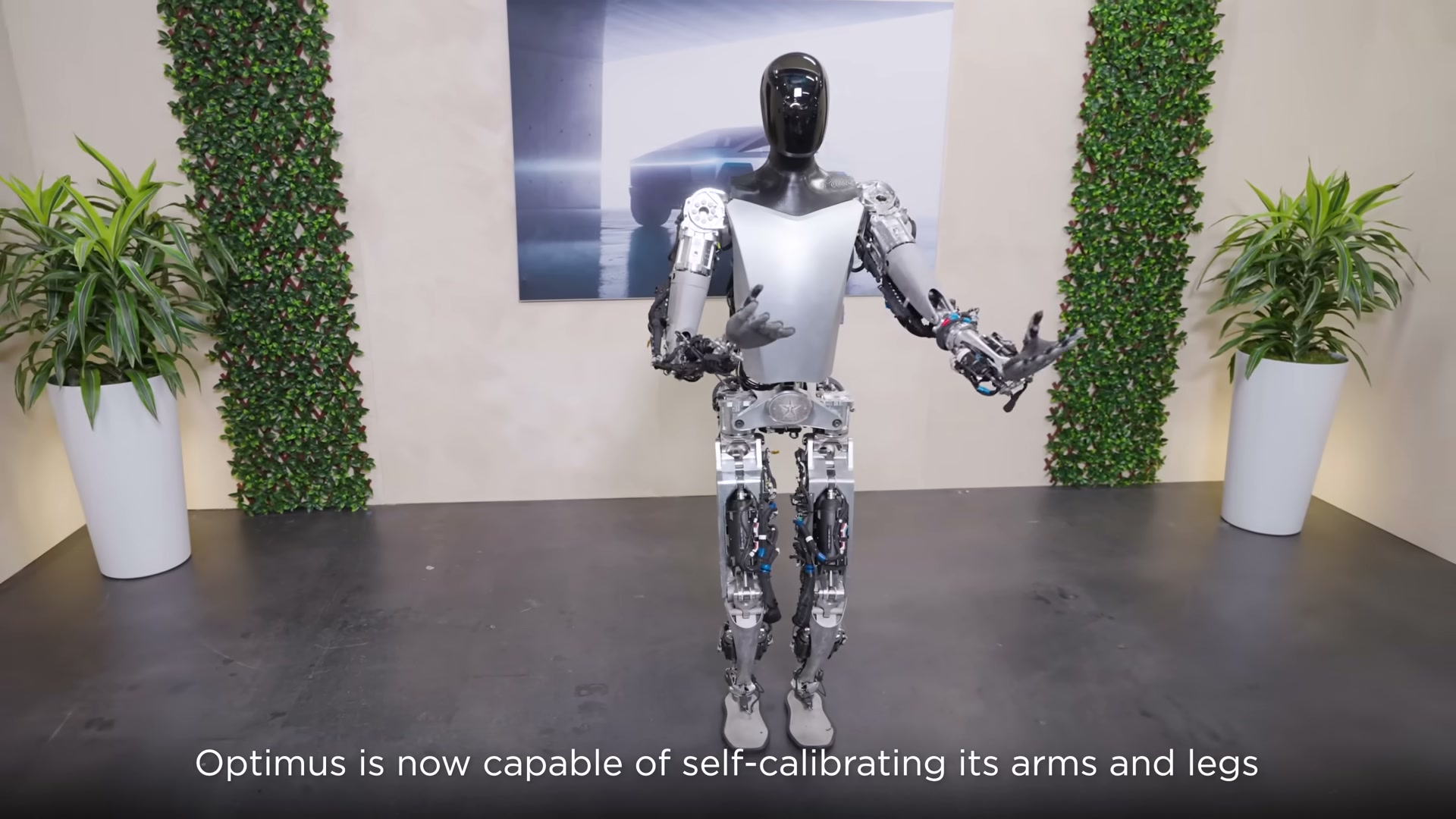 A full-body Tesla Optimus robot with exposed silver actuators and wiring standing in a studio decorated with potted plants and green ivy columns, arms slightly raised, with the caption 'Optimus is now capable of self-calibrating its arms and legs'.