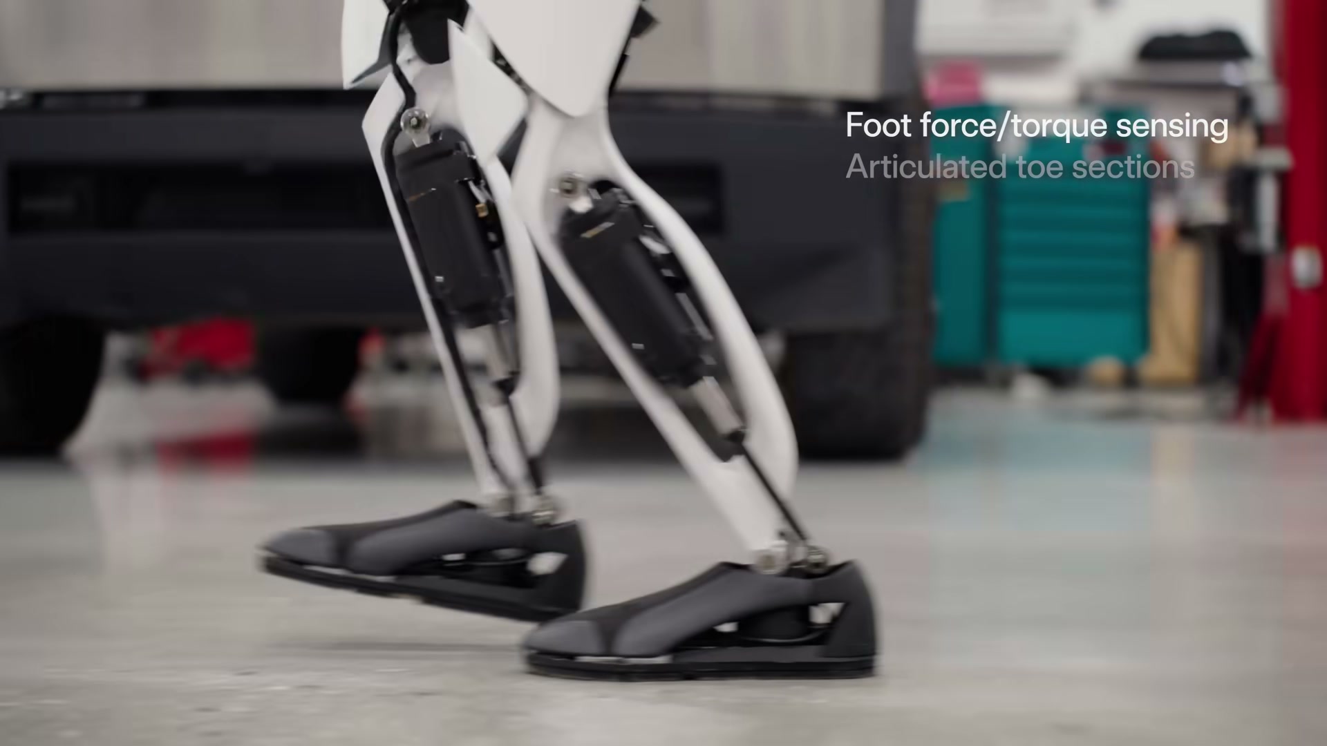 Close-up of a Tesla Optimus robot's lower legs and black articulated feet mid-step on a polished workshop floor, with the on-screen caption 'Foot force/torque sensing / Articulated toe sections' and tool chests behind.