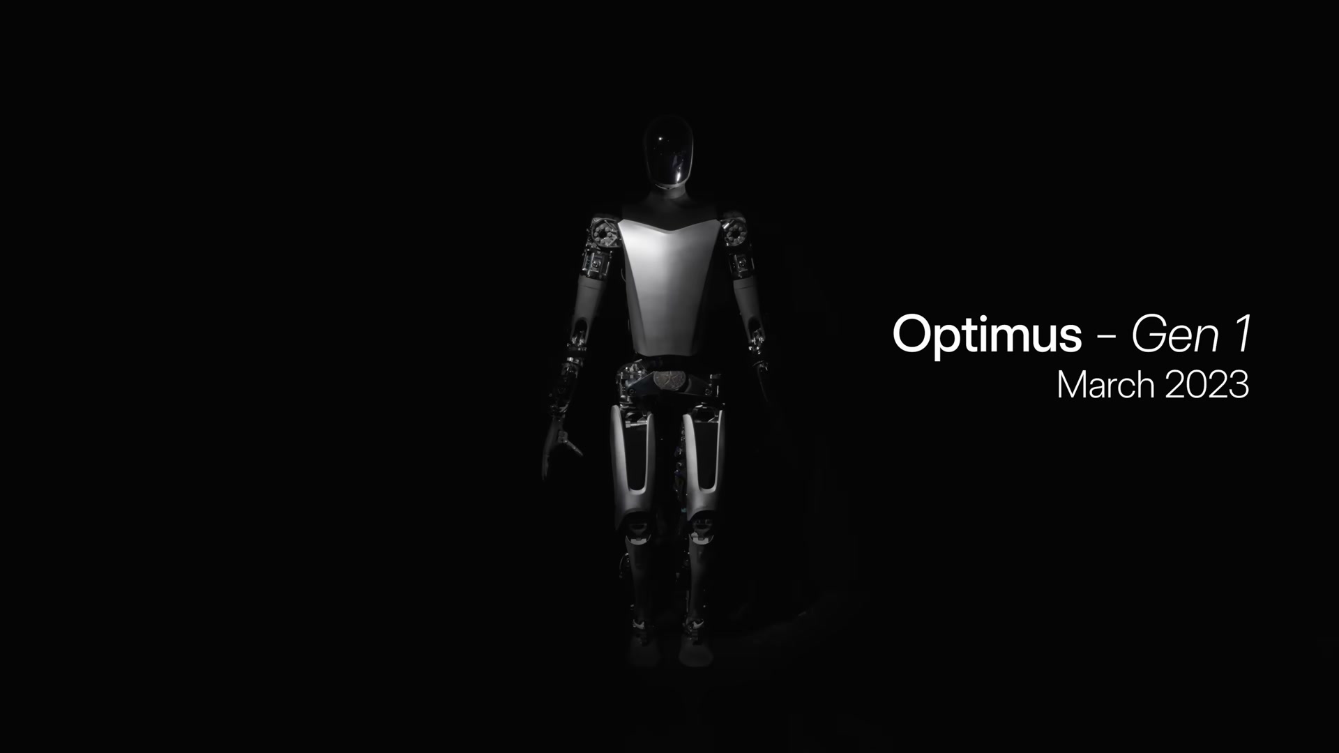 A full-body Tesla Optimus Gen 1 development robot standing on a black background with its silver-and-black body lit from the front, carrying the on-screen label 'Optimus Gen 1, March 2023'.