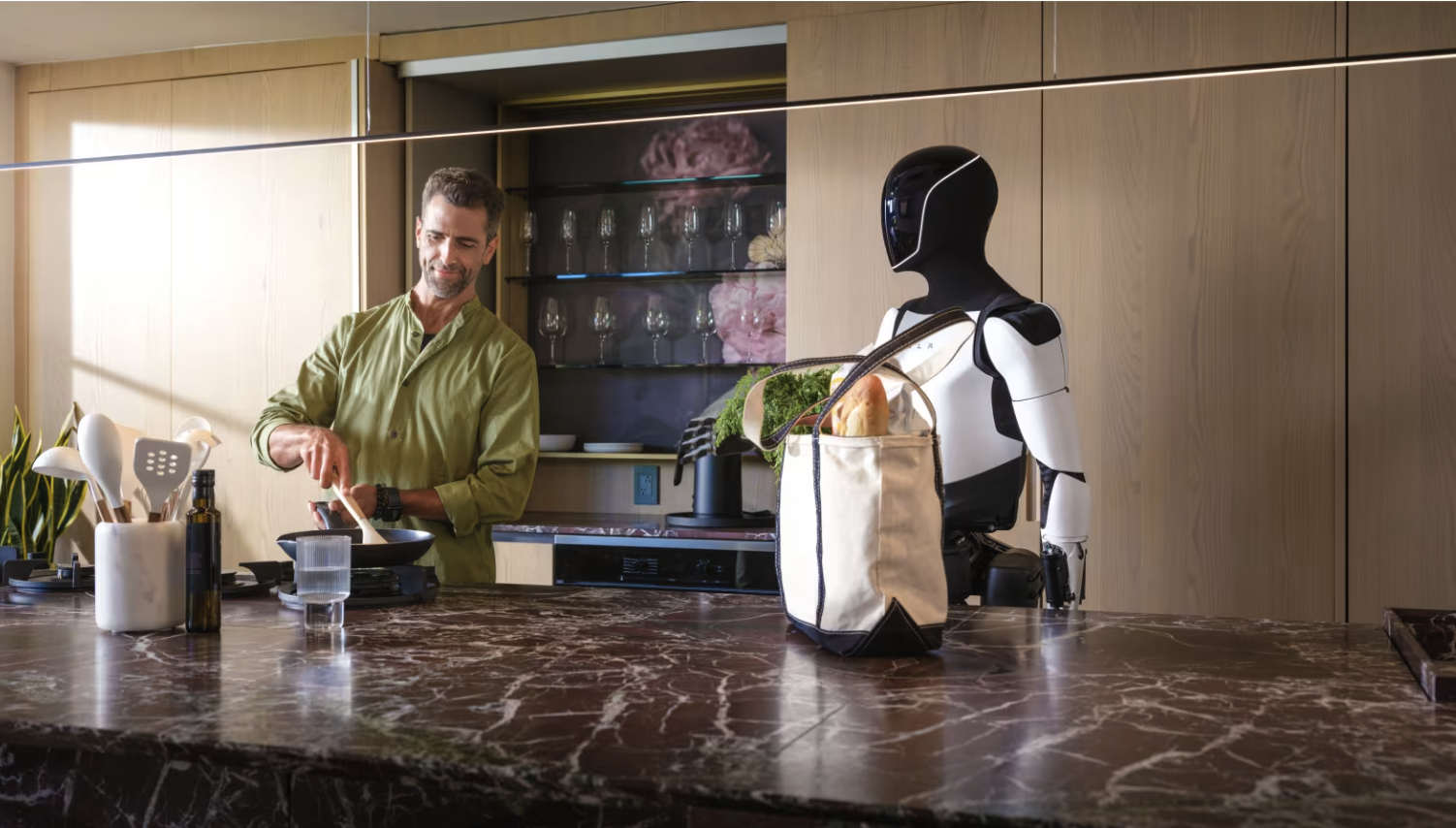 A white-and-black humanoid robot stands in a home kitchen near a tote bag of groceries while a person cooks at a counter.