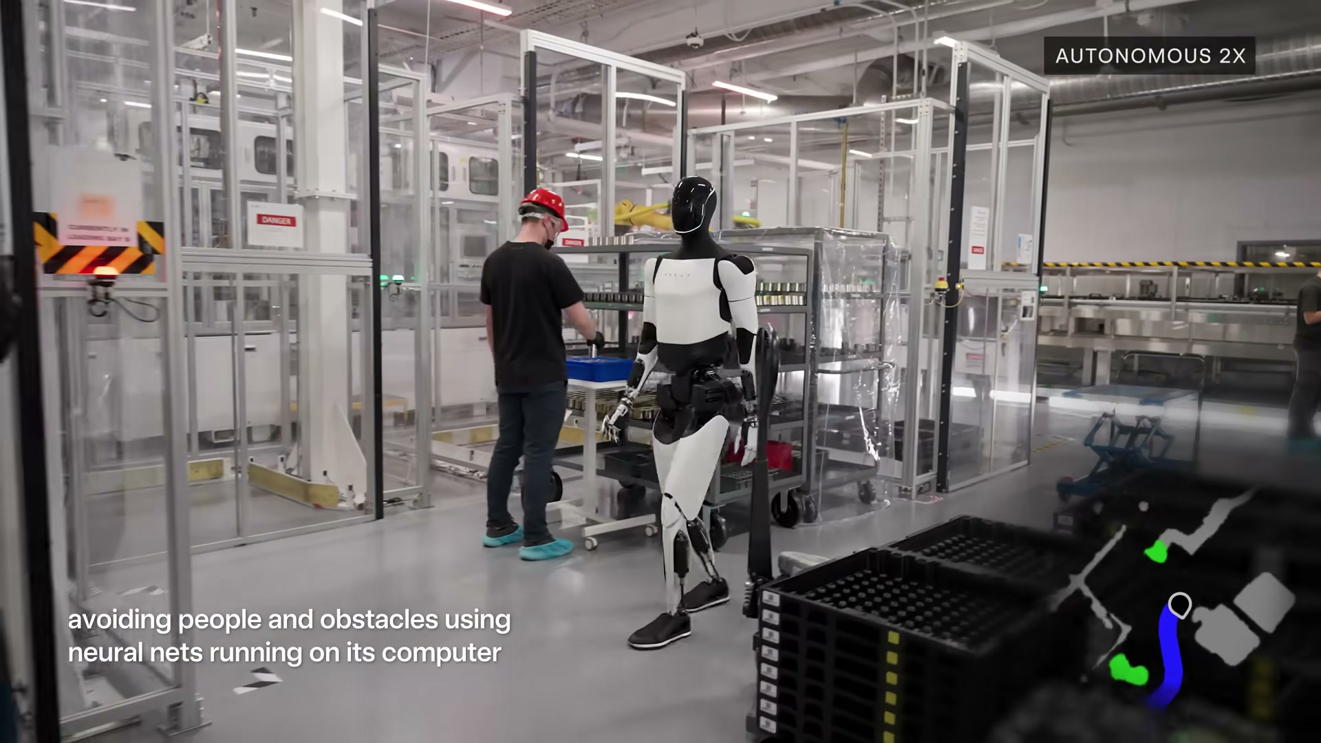 A production-design Tesla Optimus robot walking past a battery-cell workstation on a glass-walled factory floor where a human worker in a red hard hat is working, with an 'AUTONOMOUS 2X' label and a small navigation point-cloud inset in the corner.