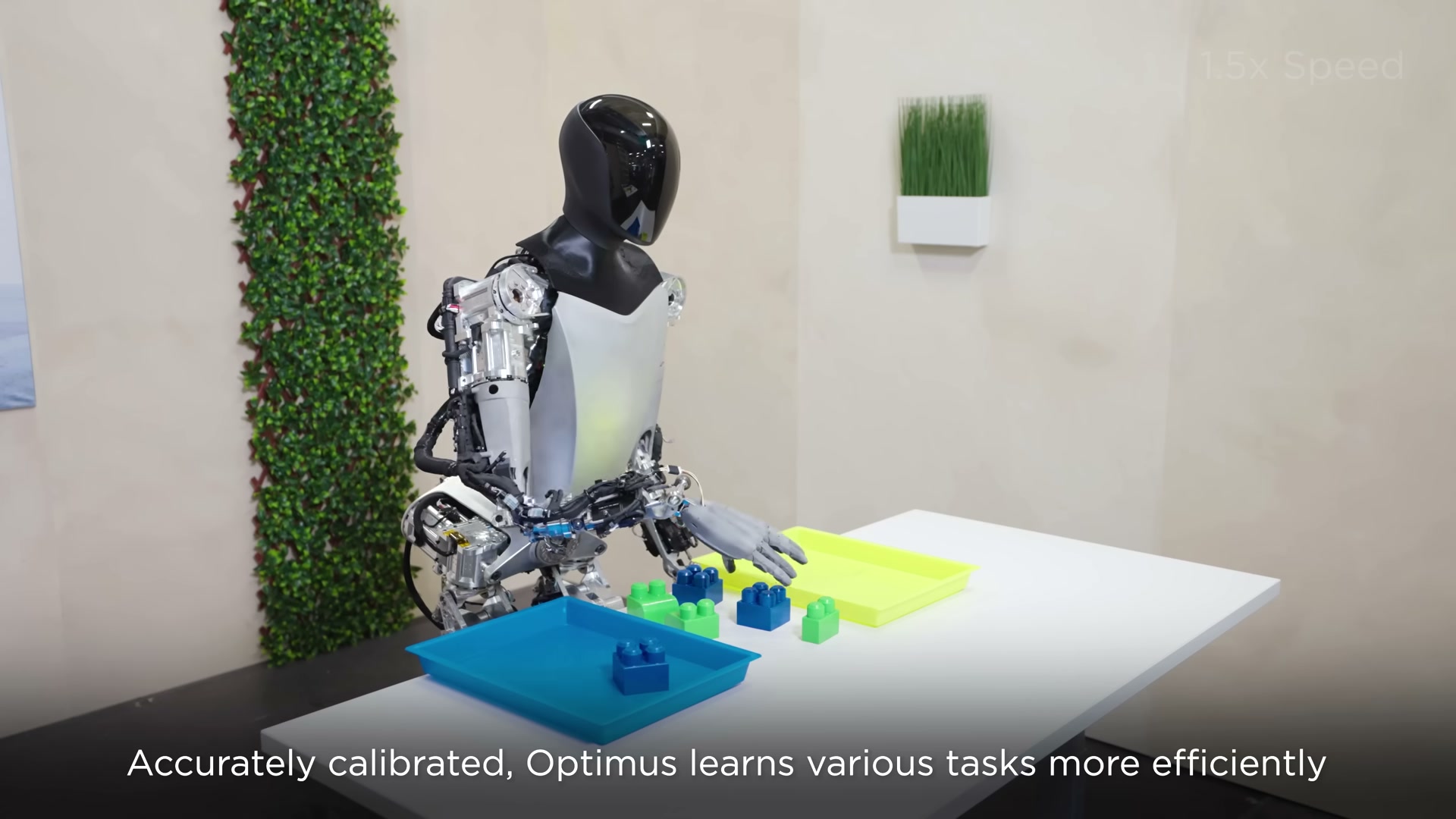 Side view of a Tesla Optimus robot with exposed silver internals reaching toward blue and green toy blocks laid out with sorting trays on a white table in a studio.