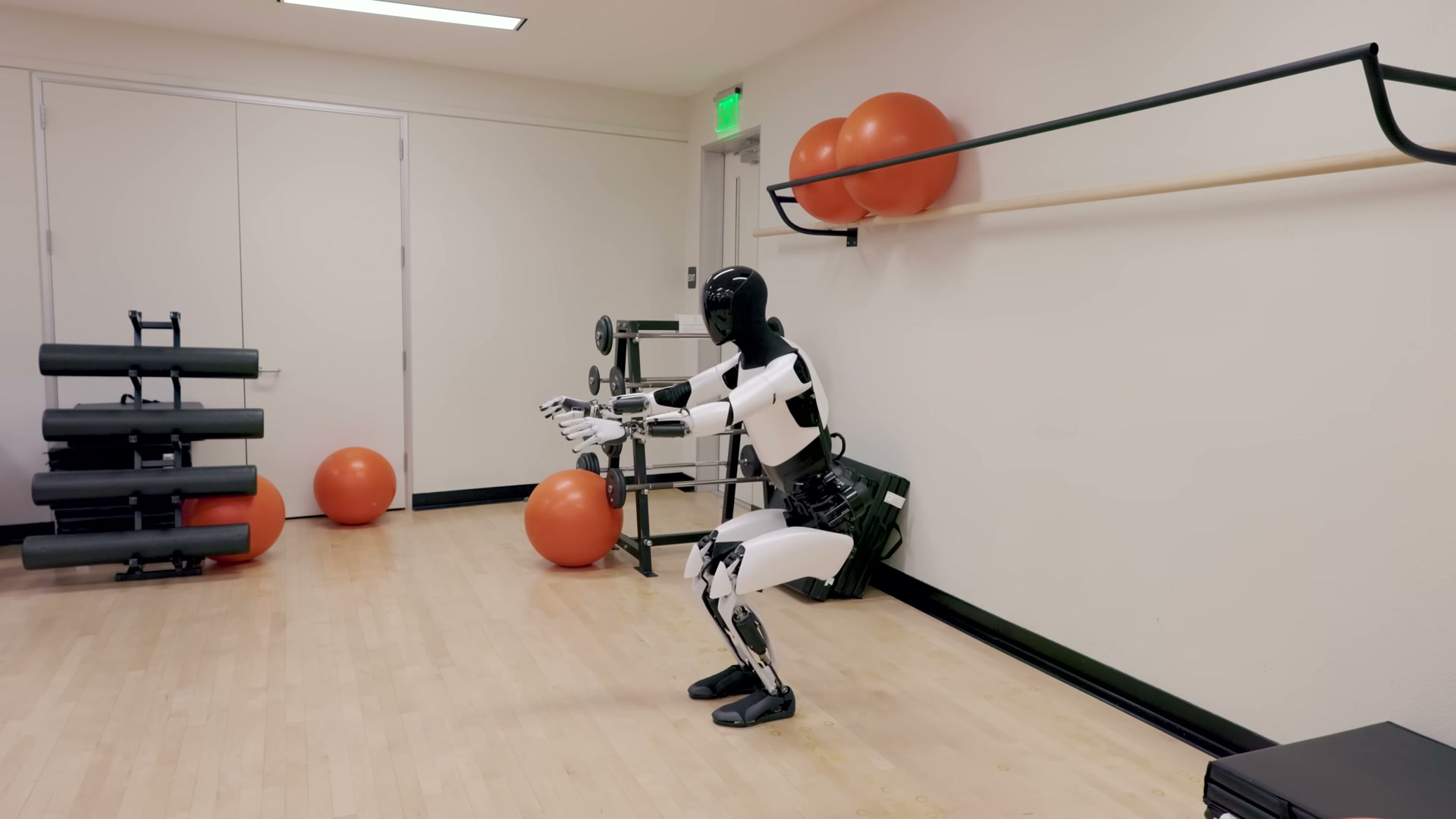 A Tesla Optimus robot performing a deep squat with arms extended forward in an exercise room, with orange stability balls, a ballet barre and a dumbbell rack in the background.