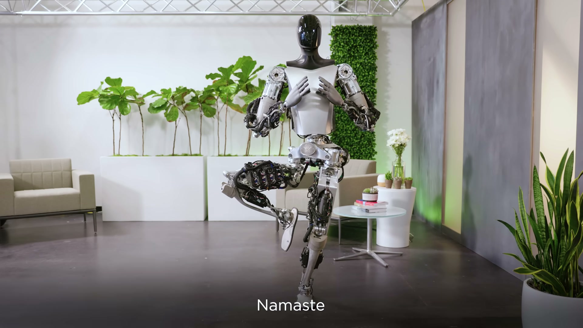 A Tesla Optimus robot balancing on one leg in a tree-pose yoga stance with its hands pressed together at its chest, in a living-room-style studio with potted plants, a sofa and a coffee table, captioned 'Namaste'.