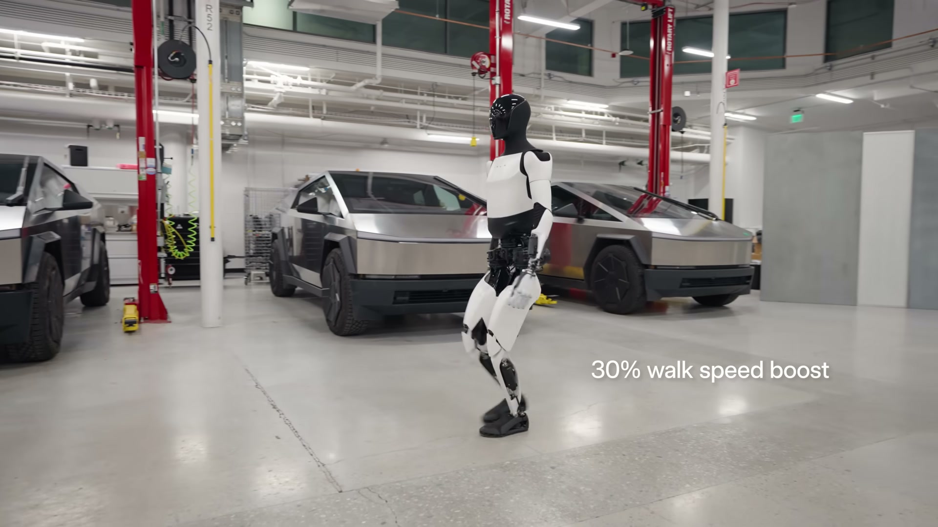 A Tesla Optimus (Gen 2) robot walking mid-stride across a Tesla service garage with two stainless Cybertrucks on red vehicle lifts behind, and the on-screen caption '30% walk speed boost'.