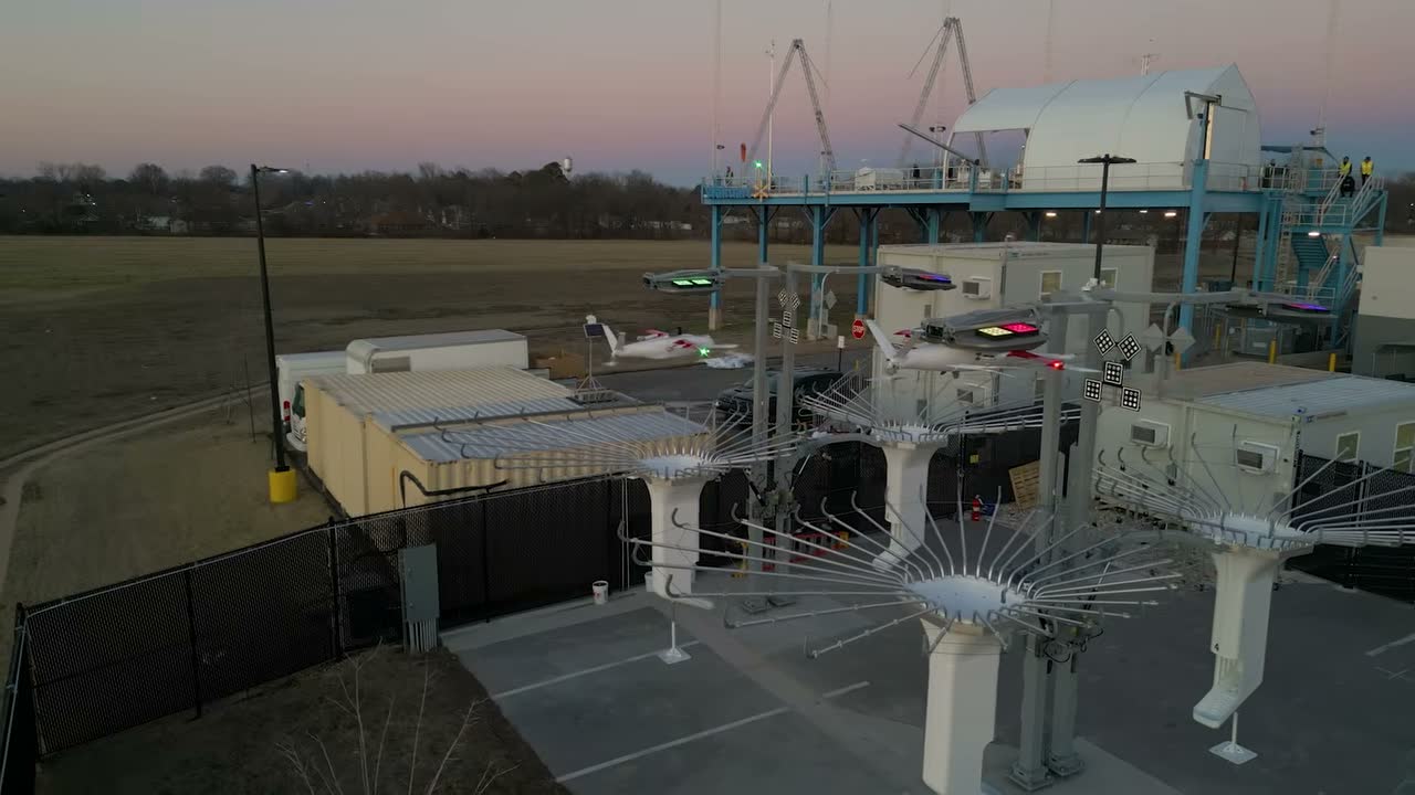 Zipline Platform 2 delivery droids docked at an outdoor loading station at dusk, with one droid airborne above the dock structure.